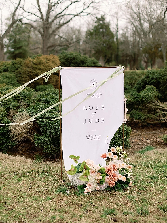 Wedding welcome sign with a fabric wedding welcome sign banner on a metal frame, green ribbons and floral base on a lawn by shrubs