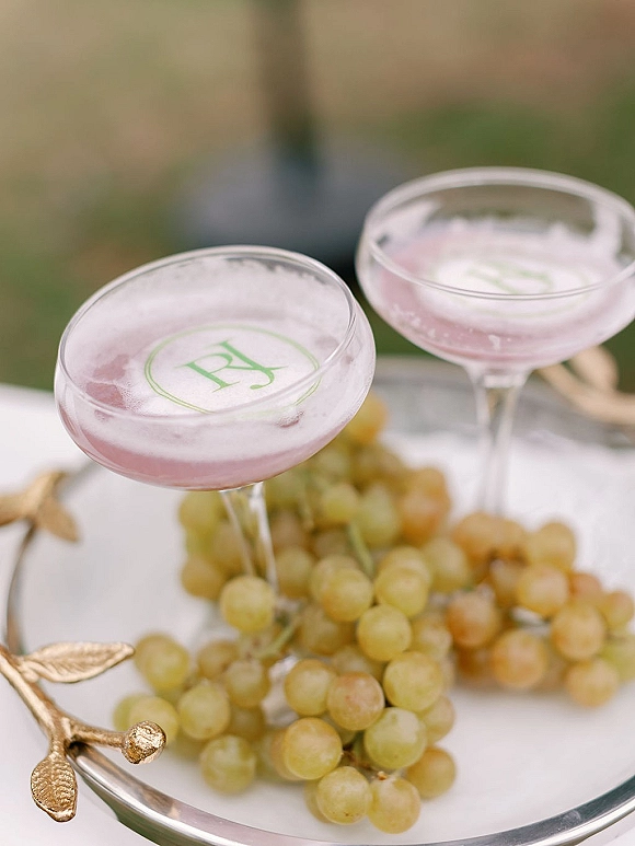 Signature cocktail in coupe glasses, a pink foam-topped wedding signature drink with monogram garnish on tray with grapes, greenery blurred behind