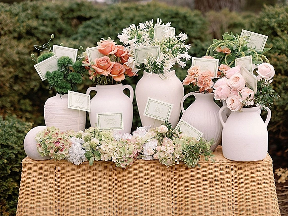 Wedding escort cards on a woven tablecloth tucked into ceramic vases with roses, carnations, and hydrangeas, set against garden shrubs
