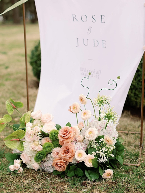 Wedding welcome sign with a fabric wedding welcome sign on a metal frame, styled with blush roses, hydrangea and greenery on a lawn