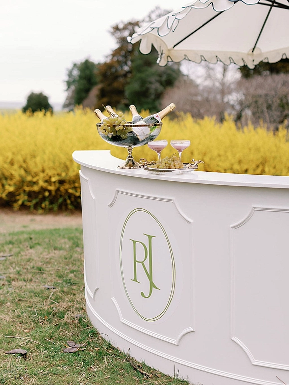 Champagne bar setup with outdoor champagne bar on a white portable counter, coupe glasses and grapes beneath a scalloped umbrella on a grassy lawn