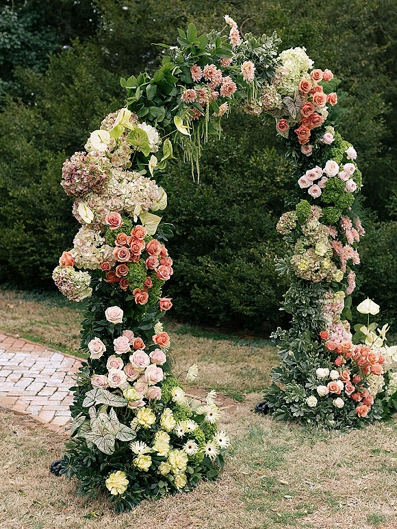 Wedding floral arch with roses and hydrangeas in an asymmetrical circle design, set on a garden lawn beside a brick pathway