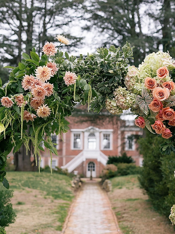 Floral wedding arch with outdoor ceremony arch flowers, greenery garland and blush dahlias with hanging amaranthus on an estate lawn near a brick house