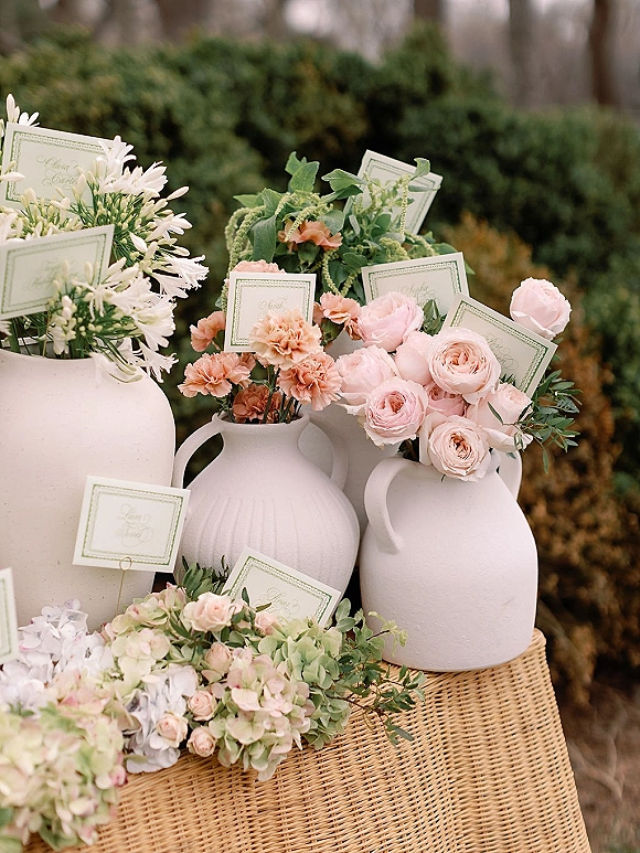 Escort card display with printed place cards nestled in white ceramic vases of blush and white flowers on a wicker pedestal outdoors