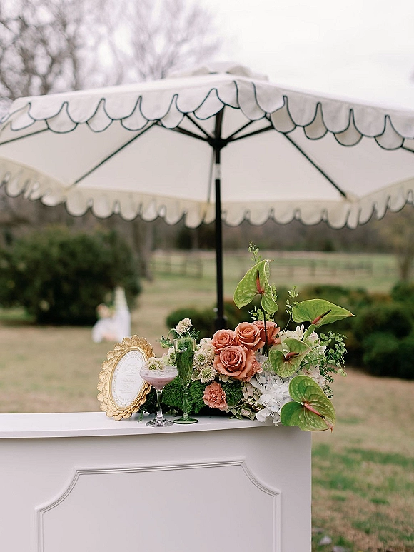Cocktail bar decor with a white portable bar under a patio umbrella, lush anthurium and rose florals, gold frame, and green goblets on a lawn