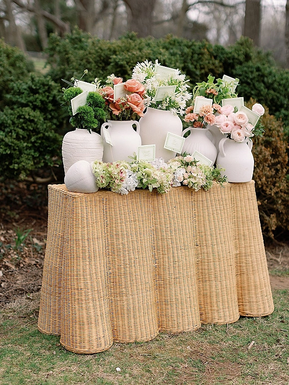 Wedding escort display with place cards tucked into flowers atop rattan pedestal tables, framed by hedge greenery and lawn grass outdoors