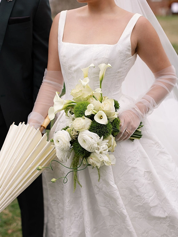 Bridal bouquet of white calla lilies and roses held by a bride in sheer gloves, with groom in tuxedo on an outdoor lawn with blurred guests