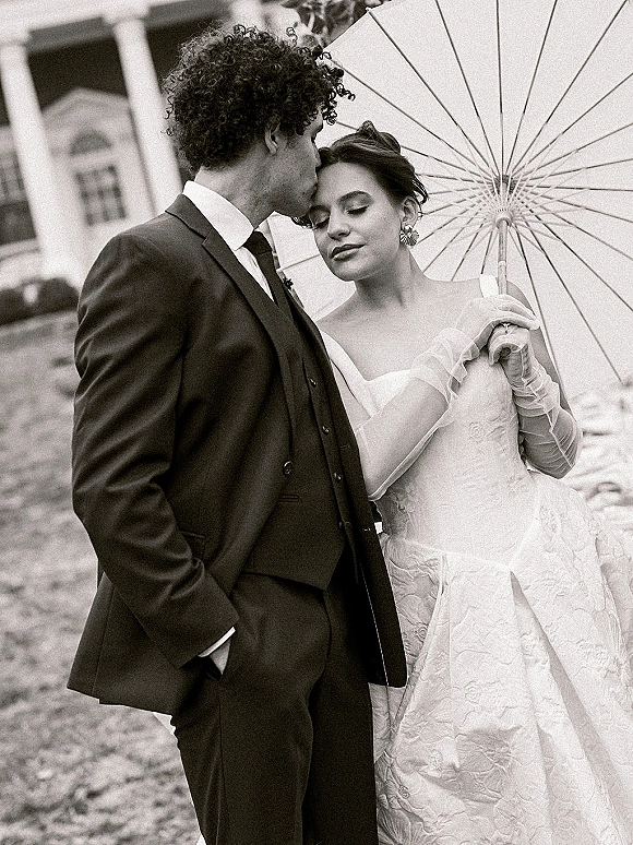 Couple portrait of bride and groom under a parasol, groom kissing her forehead, with veil and gloves on an estate lawn backdrop