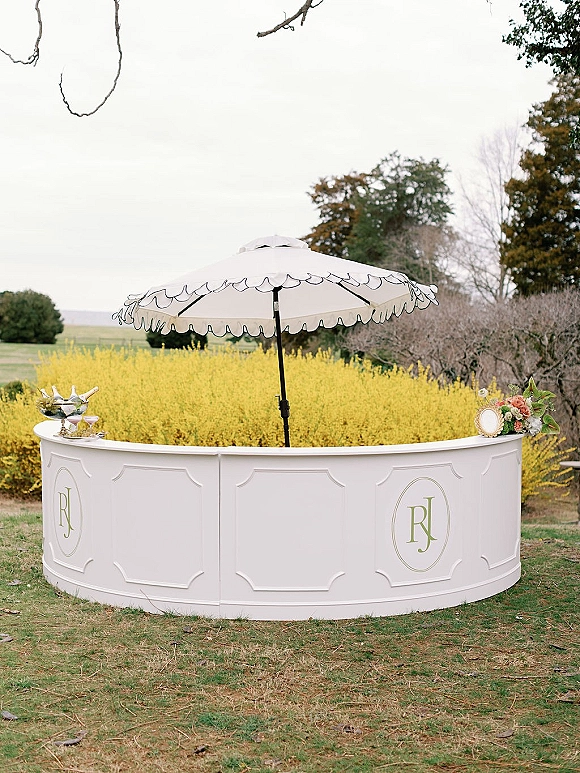 Wedding bar setup with a white outdoor bar under a scalloped parasol, champagne bucket and glassware on a grassy lawn in the countryside