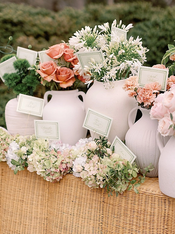 Escort card display with wedding escort cards tucked among peach roses and hydrangeas in large white ceramic vases on a woven runner, garden hedges behind