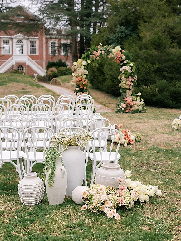 Ceremony setup with white ceremony chairs facing a peach-and-white floral arch with greenery, set on a garden lawn by a brick building