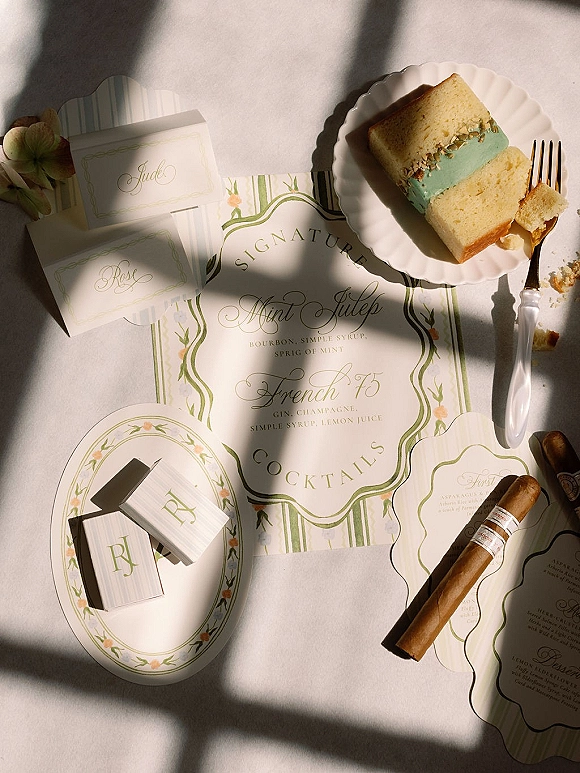 Wedding menu card with cocktail menu sign, place cards, monogram matchbooks, cake slice and gold fork on a sunlit white tablecloth