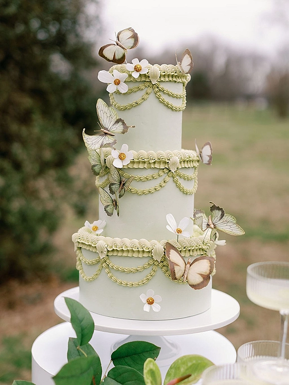 Wedding cake with butterfly cake topper, sugar flowers, and piped frosting swags on a stand at an outdoor lawn dessert table
