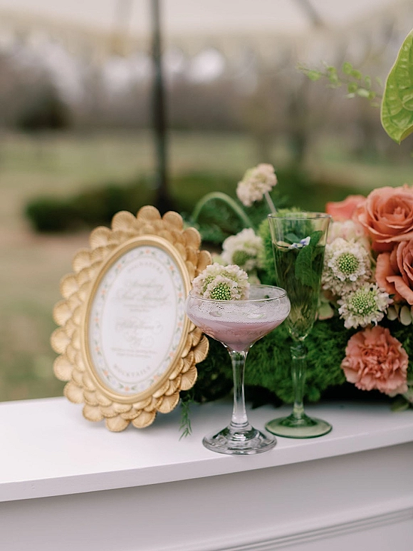 Wedding cocktail display with signature wedding cocktails in coupe and flute beside a gold-framed menu and peach rose florals on a lawn table
