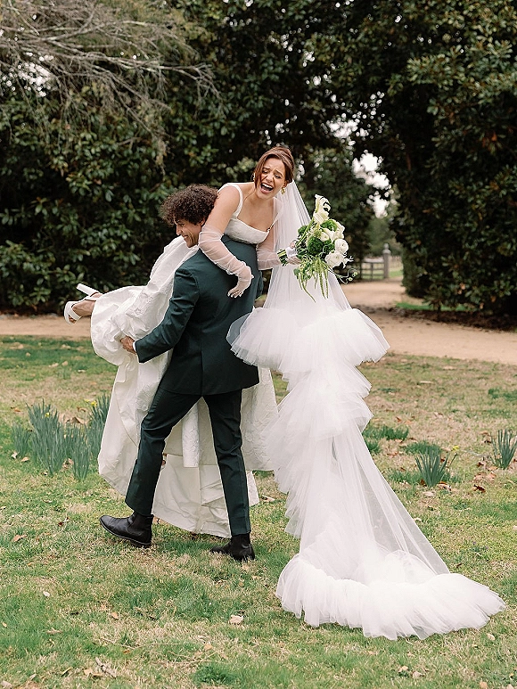 Couple portrait of groom carrying bride over his shoulder, her long veil trailing and bouquet in hand on a garden path
