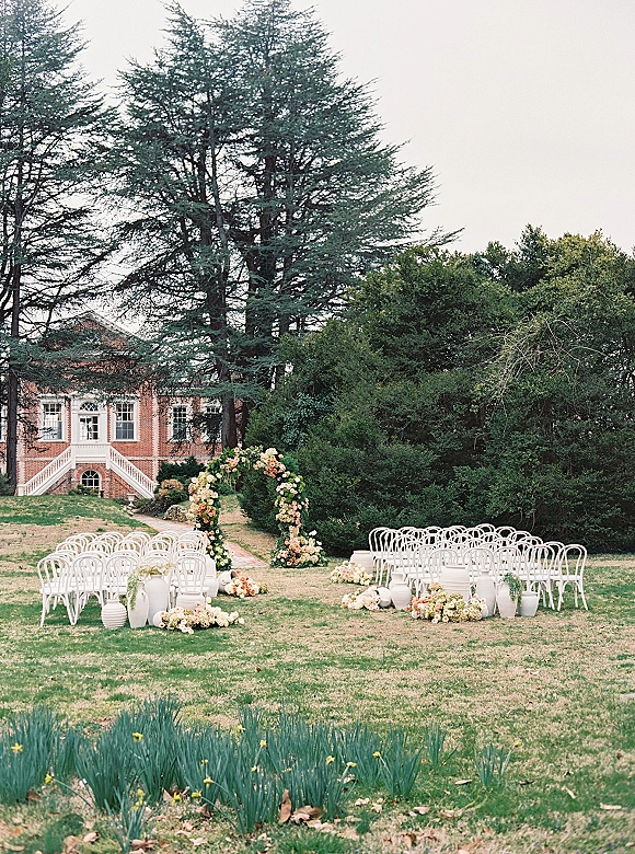 Outdoor ceremony setup with a floral arch and aisle florals, white bentwood chairs on a lawn before a brick manor house, framed by tall trees