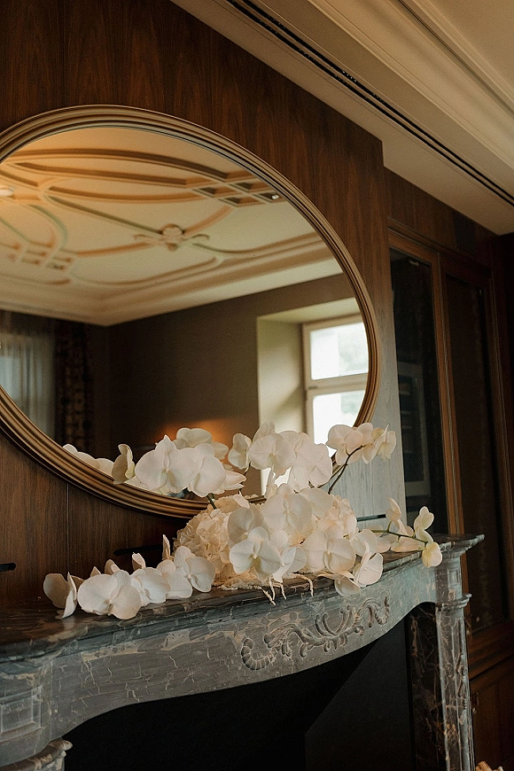 Wedding mantel decor with fireplace wedding decor featuring white orchids on a stone mantel beneath an oval mirror on a wood-paneled wall