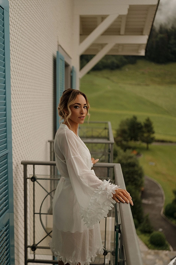 Bridal portrait of a bride on balcony in a white robe with floral sleeve appliqué, earrings and updo, looking over shoulder by blue shutters.