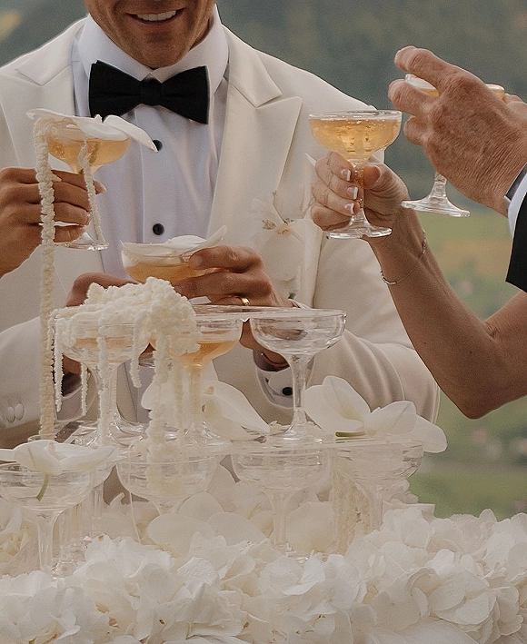 Champagne tower toast as groom in white tuxedo and black bow tie pours into coupe glasses with hydrangeas and orchids, hills blurred behind