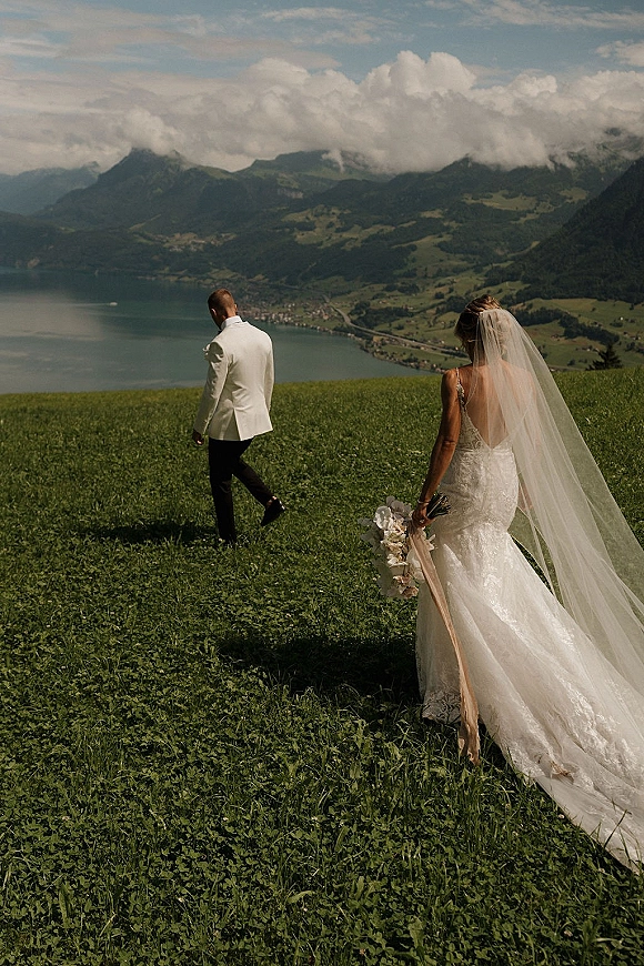 Couple portrait at a mountain wedding overlook, bride in lace dress with long veil and orchid bouquet beside groom in white jacket by a lake