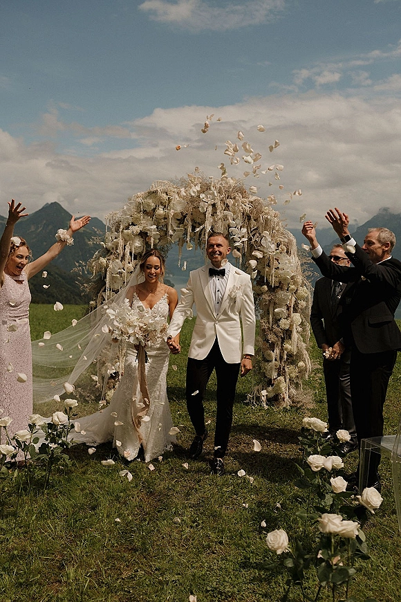 Wedding recessional with newlyweds walking aisle, bride and groom smiling under a white floral arch as rose petals fall, mountains beyond