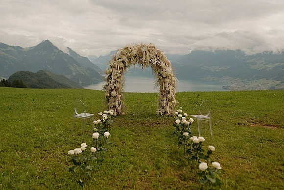 Outdoor ceremony setup with a floral arch, white flowers, and pampas grass, clear chairs and aisle blooms overlooking mountains and lake