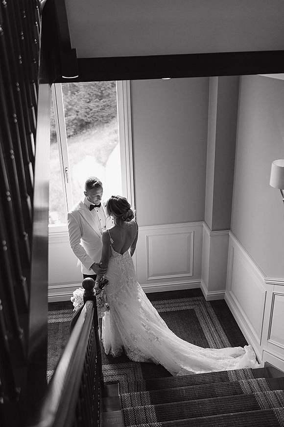 Couple portrait in a black and white wedding portrait style, bride in lace dress with train and groom in tuxedo on a staircase by a window