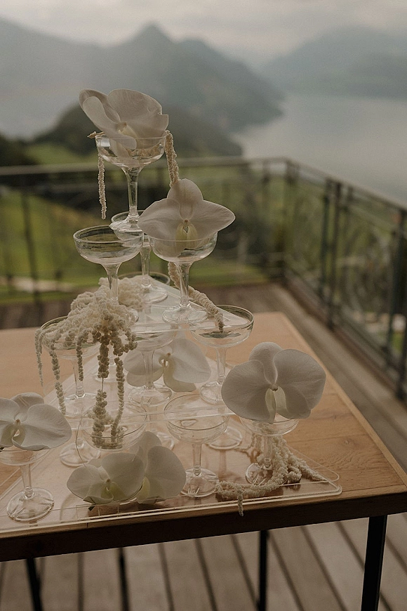 Champagne tower setup with coupe glasses and white orchid accents on a tray, set on an outdoor balcony with mountain and lake views