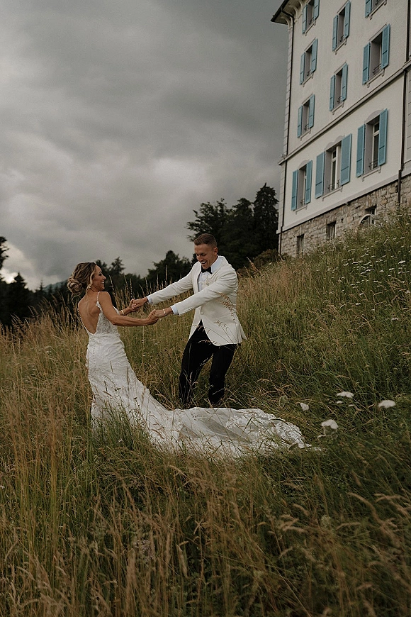 Couple portrait of bride and groom holding hands as she twirls, lace train flowing in a wildflower meadow by a stone building under clouds