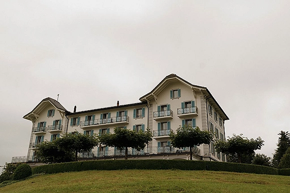 Wedding venue exterior with balconies and shuttered windows, wrought iron railings, framed by trees and hedges on a grassy hill under overcast sky