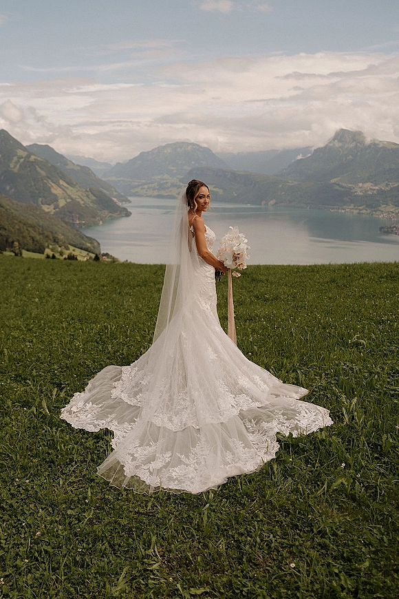 Bridal portrait of a bride in a lace wedding dress with long veil, holding an orchid bouquet in a mountain lake meadow under cloudy sky