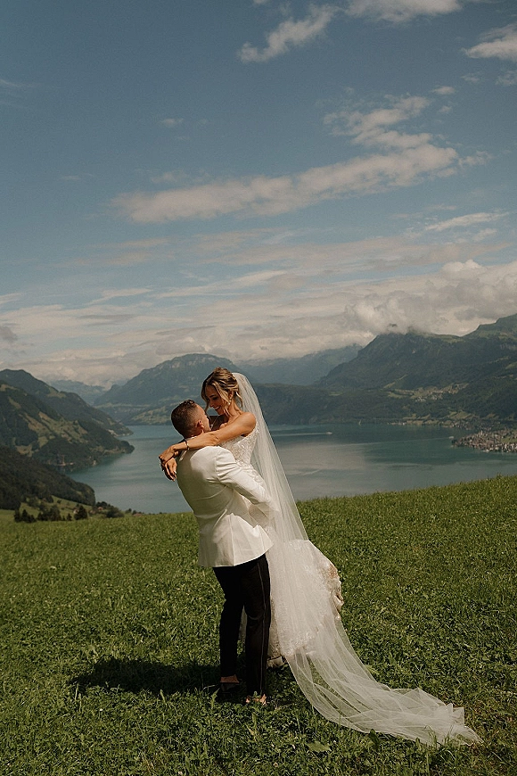 Couple portrait of groom lifting bride in a strapless lace gown, long veil flowing on a grassy hill above a lake and mountains