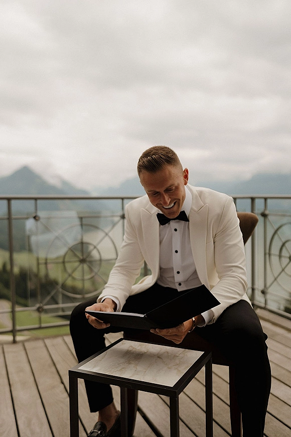 Groom portrait in a white tuxedo jacket and black bow tie, seated reading a vow book on an outdoor terrace with mountains behind