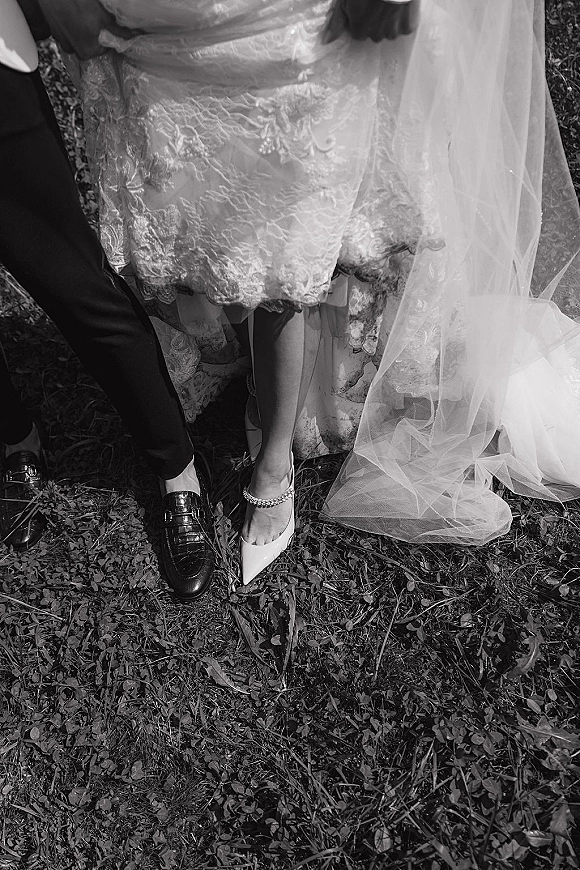 Wedding shoes detail of bride and groom shoes on grass with fallen leaves, lace dress hem and tulle veil framing her heels and ankle bracelet
