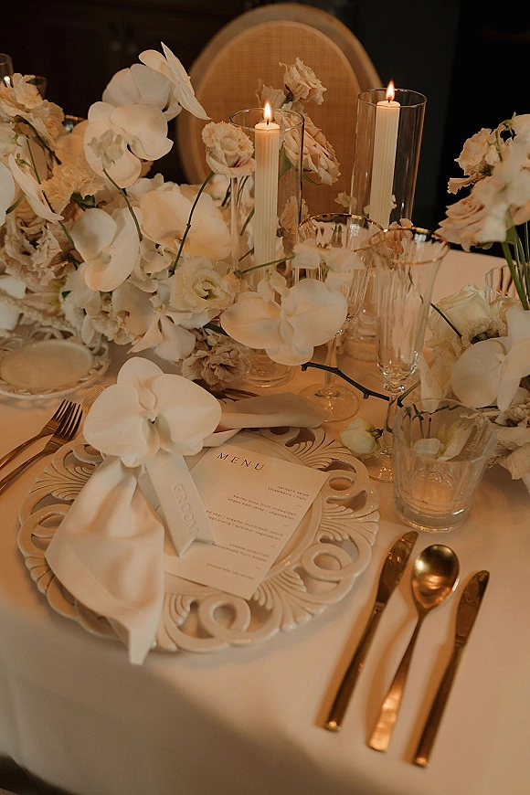 Reception tablescape with wedding table setting, white florals and orchid centerpiece, taper candles in glass hurricanes, gold flatware and menu cards in dim light