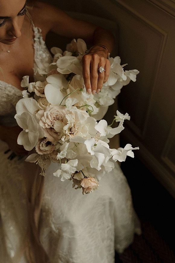 Bridal bouquet of white orchids and blush roses held against a lace wedding dress, with engagement ring and pearl necklace on wooden stairs