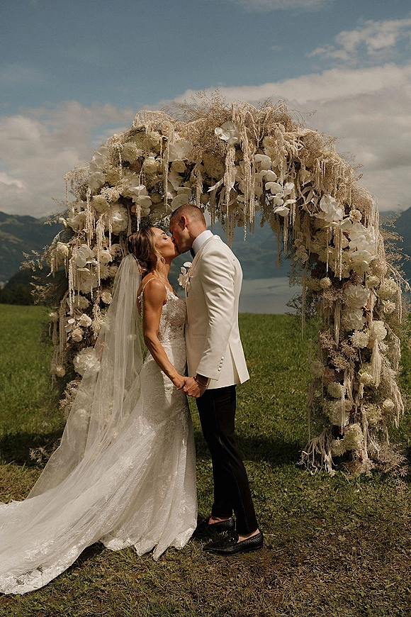 Wedding kiss portrait of bride and groom kissing under a white floral arch, her veil flowing, with mountains and lake behind under clouds
