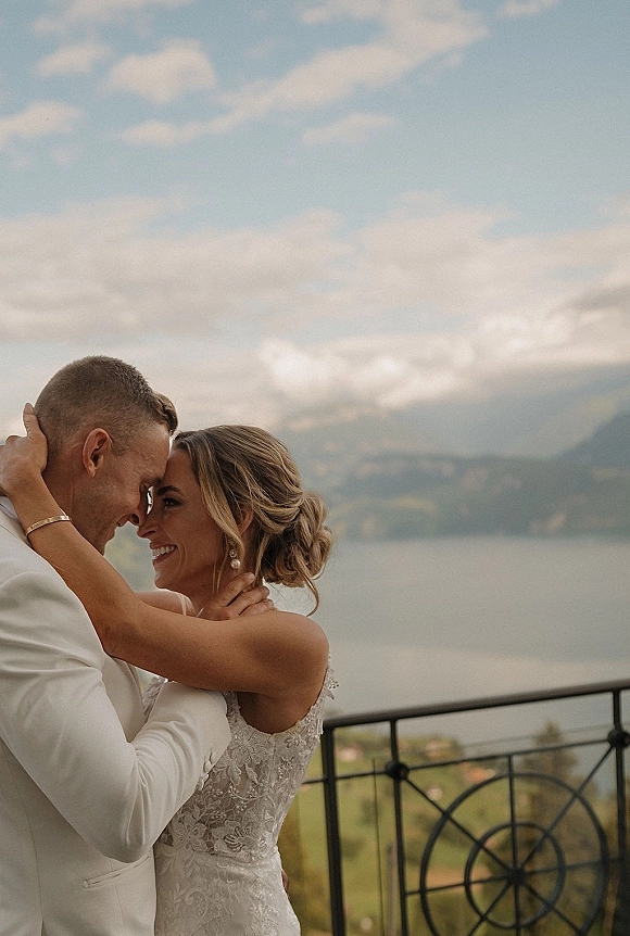 Couple portrait of bride and groom forehead touch, embracing by a balcony railing with mountains and valley under a cloudy sky