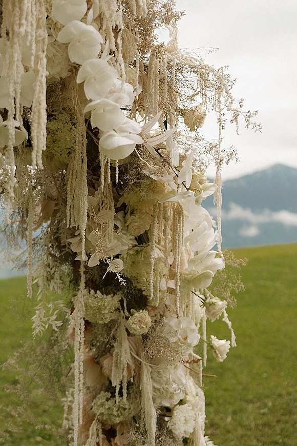 Wedding ceremony flowers cascade with white orchids and roses on a wooden post, with airy greenery against a mountain meadow backdrop