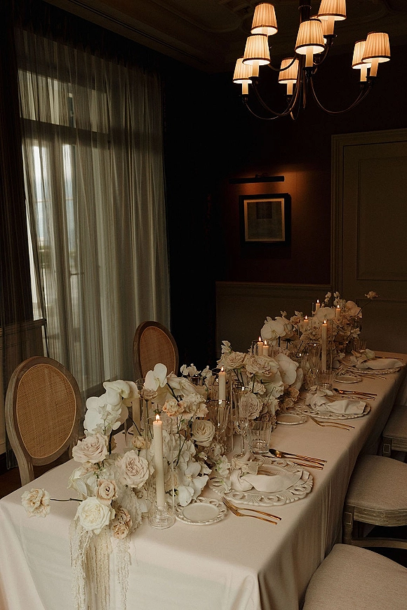 Reception tablescape with long head table decor, white floral centerpiece and taper candles on a white tablecloth beneath a chandelier