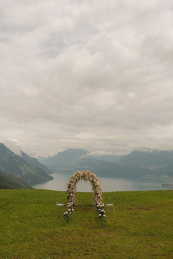 Ceremony arch with white floral cascade and hanging strands, set on a grassy hill with clear chairs overlooking misty lake and mountains