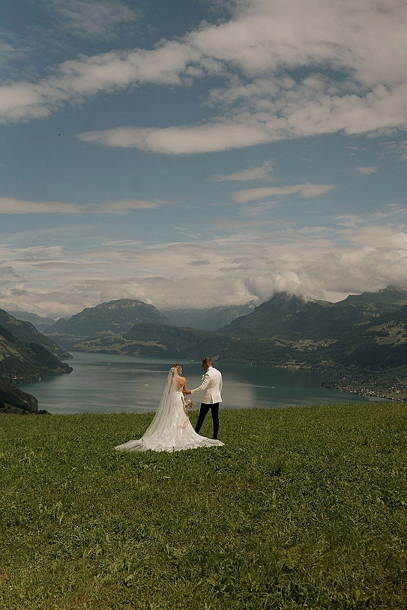 Couple portrait in mountain wedding photos, holding hands as the bride’s long veil trails beside a lake under a cloudy sky