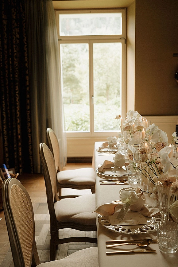Reception tablescape with long head table decor, white floral centerpieces and taper candles on white linens by a window with sheer curtains