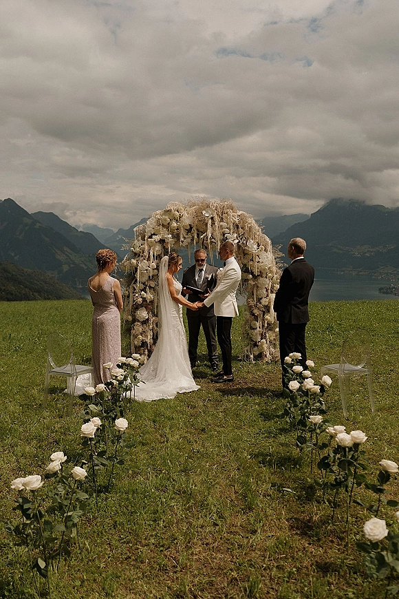 Wedding ceremony with bride and groom exchanging vows under a white rose floral arch, with clear chairs beside a mountain lake meadow backdrop