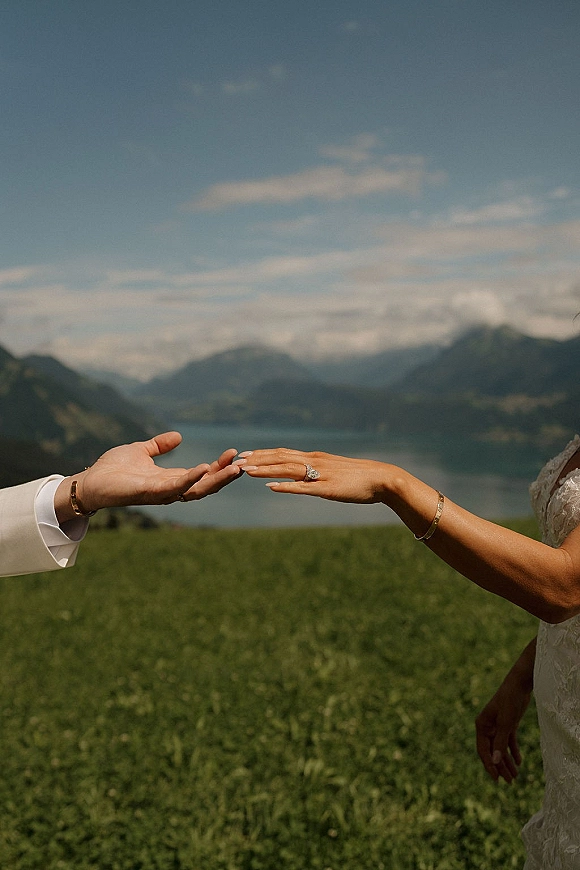 Wedding ring exchange as groom slides a band onto bride’s manicured hand, engagement ring sparkling, with mountains and lake behind