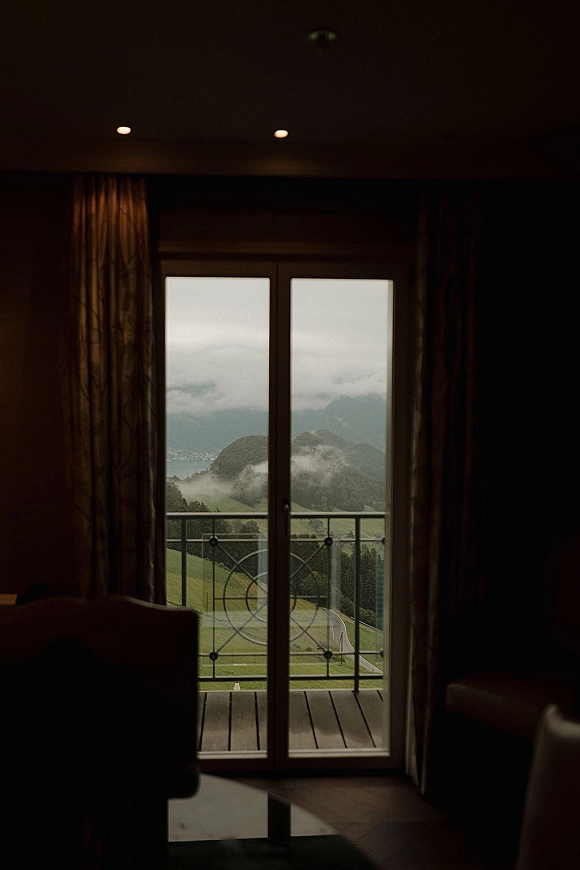 Hotel room view with mountain view balcony through a sliding glass door, curtains framing a misty lake and hillside under low clouds