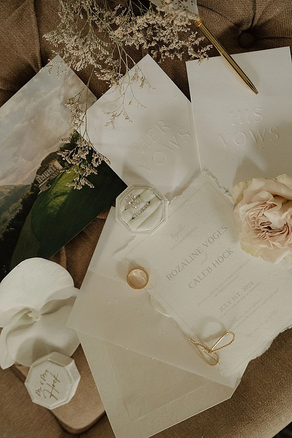 Invitation suite wedding invitation flat lay on linen fabric with embossed vow booklets, wax seal envelope, blush rose, gold pen, and rings