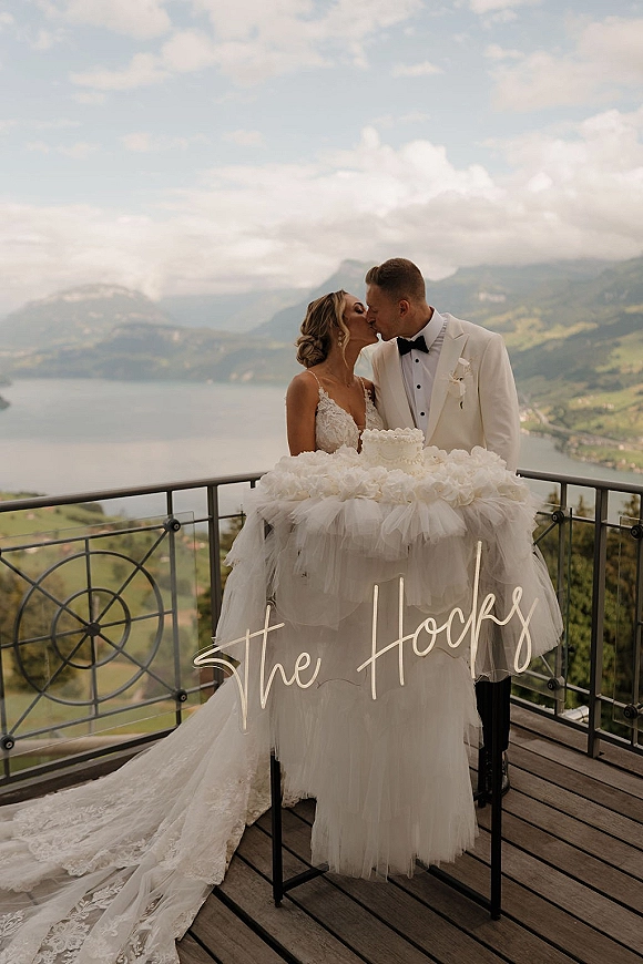 Wedding kiss portrait of bride and groom kissing by a cake table with lace gown and white tux, on a balcony overlooking mountains and a lake