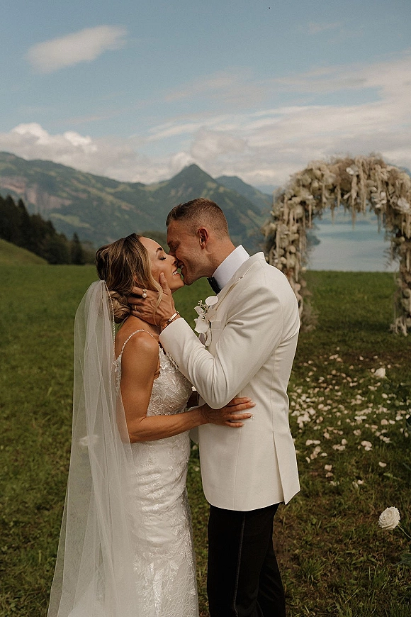 Wedding kiss as bride and groom share a close embrace under a dried floral arch, mountains and lake behind, veil and petals in view