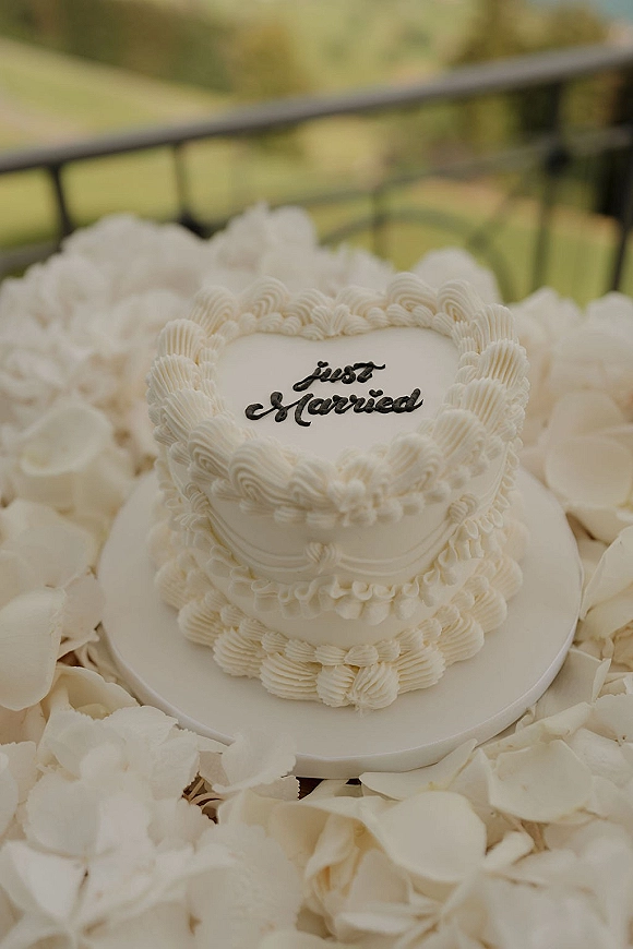 Wedding cake with white buttercream wedding cake piping and black script lettering on a stand, set outdoors with greenery and scattered petals